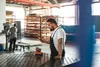 Two men working in a factory setting. One man in the foreground is looking at his phone while leaning on a workbench. Another man is in the background operating machinery. Produktbild-Vorschau