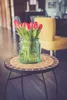 A small round table with a mosaic top holds a glass vase filled with pink tulips. A yellow chair is visible in the background. Produktbild-Vorschau