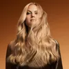 A woman with long blonde hair is shown from the chest up, with her hair flowing around her face. She is wearing a shimmering gold top. Produktbild-Vorschau