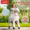 A young girl is riding a plush unicorn toy in an outdoor setting with greenery in the background. Produktbild-Vorschau
