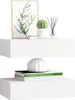 Two white floating shelves are displayed against a white background. The top shelf holds a framed picture of greenery, a small potted cactus, a book, and a diffuser with reeds. The bottom shelf displays two books and a white decorative vase. Produktbild-Vorschau