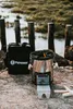 A Petromax rocket stove and its carrying bag are placed on a rocky surface near a body of water with wooden posts in the background. Produktbild-Vorschau