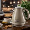 A vintage-style electric kettle is displayed on a wooden table, alongside a cup of tea, a slice of pie, and some cookies. The background shows a softly lit kitchen area. Anteprima dell’immagine prodotto