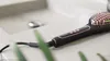 A close-up shot of a black and rose gold electric hair straightening brush resting on a light gray countertop next to a sink. Produktbild-Vorschau