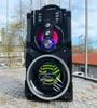A portable Bluetooth speaker is placed outdoors on a metal surface, with buildings and trees in the background. Produktbild-Vorschau