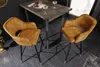 Two mustard yellow velvet bar stools with quilted detailing and gold accents are placed around a dark gray table with champagne glasses on it. Produktbild-Vorschau