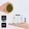 A hand holds a glass jar with a golden lid, and another hand holds a glass jar with dimensions indicated. Icons showing weight and diameter are also present. Voir les images du produit