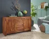 A wooden sideboard with drawers and doors, decorated with vases, a globe, and books, in a living room setting with a sofa and plants. Produktbild-Vorschau