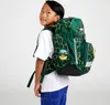 A young boy wearing a white t-shirt and blue pants is shown from the side, wearing a green backpack with a yellow geometric pattern. Produktbild-Vorschau