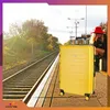 A woman in a red hat sits on a train platform next to a yellow suitcase, waiting for a train. Anteprima dell’immagine prodotto