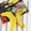 A yellow banana-shaped storage box is open, with a banana inside, placed on top of a magazine. Produktbild-Vorschau