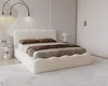 A bedroom scene featuring a bed with a textured headboard and frame, dressed with bedding and pillows. A bedside table with decorative items and a lamp are also visible. Náhled obrázku produktu