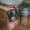 A person's hands are shown opening a stainless steel container, possibly a Schinkenkocher, on a wooden countertop. In the background, a clear container filled with microgreens is visible. Produktbild-Vorschau