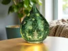 A decorative glass teardrop-shaped lamp with a Christmas theme, placed on a wooden table. Produktbild-Vorschau