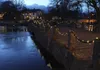 A stone bridge decorated with fairy lights at dusk, with a building in the background. Produktbild-Vorschau