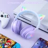 A pair of purple gaming headphones with cat ears is placed on a light purple surface next to a white gaming console and a white controller. A smartphone and a Nintendo Switch are also visible in the foreground. Podgląd zdjęcia produktu
