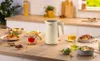 A Bosch electric kettle is displayed on a kitchen counter surrounded by various food and drink items, suggesting a breakfast or brunch setting. Produktbild-Vorschau