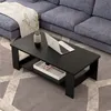 A black coffee table with a shelf is placed on a gray patterned rug in front of a blue sectional sofa. A laptop, a vase with greenery, and a white tray with items are on the table. Produktbild-Vorschau
