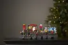 A Christmas-themed train decoration with Santa and a nutcracker figure, placed on a dark surface with a blurred Christmas tree in the background. Podgląd zdjęcia produktu