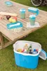 A picnic setup with food and drinks on a wooden table and a cooler on the grass. Podgląd zdjęcia produktu