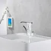 A modern bathroom sink with a waterfall faucet running water into it. A small framed picture and a branch with white flowers are on the wall behind the sink. Podgląd zdjęcia produktu