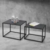 Two black metal frame tables of different sizes are placed on a concrete floor in front of a textured gray wall. The larger table on the left has three glass candle holders with candles on top. The smaller table on the right has a book on it. Náhľad obrázku produktu