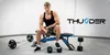 A man sitting on a weight bench with various weights and equipment around him. Náhľad obrázku produktu