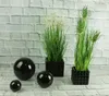 Three black decorative spheres and two potted artificial plants are arranged on a surface in front of a white brick wall. Produktbild-Vorschau
