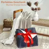 A dark blue sherpa blanket is neatly folded and tied with a red ribbon, presented as a gift. Another white and gray sherpa blanket is draped over a wooden chair in the background, with a woven basket and books nearby. A white vase with dried flowers sits o Náhľad obrázku produktu