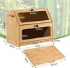 A bamboo bread box with a cutting board accessory, shown with dimensions indicated by text overlays. The background features a blurred image of bamboo stalks. Produktbild-Vorschau