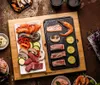 A top-down view of a Mahlzeit lavastone grill set with various ingredients arranged on a wooden board and the grill itself. Produktbild-Vorschau
