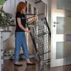 A woman is holding a foldable drying rack near a doorway, with a dog resting on a bed in the background. Produktbild-Vorschau