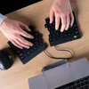 A person's hands are typing on two split ergonomic keyboards placed on a wooden desk next to a laptop and a mouse. Produktbild-Vorschau