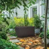 A brown storage box sits in a garden setting with fallen leaves on the ground and plants surrounding it. Podgląd zdjęcia produktu