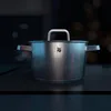 A close-up shot of a stainless steel pot with a lid handle, set against a dark, blurred background with bokeh lights. Náhľad obrázku produktu