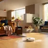 A robot vacuum cleaner is cleaning a modern living room with a sofa, fireplace, and plants. Produktbild-Vorschau