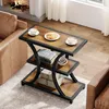 A 3-tier side table with a rustic brown wood finish and black metal frame, placed next to a brown leather sofa in a living room setting with a fireplace in the background. Produktbild-Vorschau