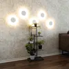 A dark gray metal plant stand with several potted plants is placed against a patterned wallpaper. Several circular lights are mounted on the wall above the plant stand. Náhľad obrázku produktu