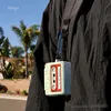 A person wearing a black jacket with a product attached to the strap, outdoors with palm trees and houses in the background. Produktbild-Vorschau