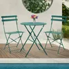 A dark green metal bistro set with a round table and two chairs is placed on a wooden deck next to a swimming pool. A vase of colorful flowers sits on the table. Produktbild-Vorschau