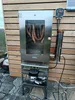 A stainless steel smoker with sausages hanging inside, placed outdoors against a wooden wall. Náhled obrázku produktu