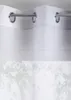 A close-up view of a white sheer curtain with grommets hanging from a curtain rod, with a blurred background of green leaves and branches. Produktbild-Vorschau
