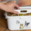 A hand is reaching into a white pet food storage container filled with pet treats. The container has cartoon stickers of a cat and a dog on it. Anteprima dell’immagine prodotto
