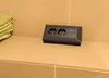 A black power strip with two outlets and two USB ports is placed on a tiled surface next to a stack of folded towels. Produktbild-Vorschau