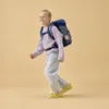 A young girl wearing a backpack walks forward against a plain background. Produktbild-Vorschau