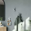 Three decorative hooks shaped like figures are mounted on a gray wall. One hook holds a pair of headphones, another holds a green jacket, and the third is empty. A mirror and a wooden cabinet are visible to the left, and a plant is on the right. Náhled obrázku produktu