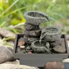 A small, stacked stone water fountain with water flowing between the stones, set in an outdoor, natural environment with greenery and rocks. Produktbild-Vorschau