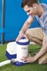 A man is kneeling next to a pool filter pump, appearing to be setting it up or checking it. The pool is visible in the background. Voir les images du produit