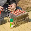 Tragbarer Grill aus Edelstahl mit Fleischspießen auf einem hellbraunen Klapptisch im Freien, neben einer Gewürzflasche und einem dunklen Kessel Anteprima dell’immagine prodotto