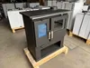 A dark gray wood-burning stove is displayed in a warehouse setting, with other similar stoves or appliances stacked in the background. Produktbild-Vorschau